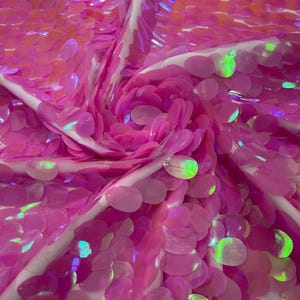 May include: A fabric material densely covered in round, bright pink sequins. The sequins have an iridescent finish that reflects light with green and yellow highlights. The fabric is gathered into a central spiral, creating a textured, layered effect.