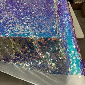 May include: A roll of iridescent sequin fabric. The fabric is a blend of blue, purple, and teal, with a shimmering, reflective quality. The sequins are small and densely packed, creating a textured surface. The fabric is on a white surface.
