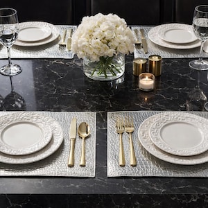 May include: Elegant table setting with white plates, gold cutlery, and crystal glassware. The table is set with silver placemats, a floral centerpiece, and candles. A sophisticated arrangement for a special occasion or dinner party.