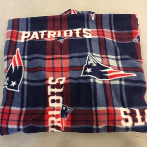 May include: A blue, red, and white fleece blanket with a plaid pattern and the word "Patriots" printed on it.