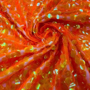 May include: A vibrant orange fabric densely covered in circular, iridescent sequins. The material is gathered in a central swirl, creating a textured, reflective surface where the light catches the shiny, metallic-like finish of each individual disc.
