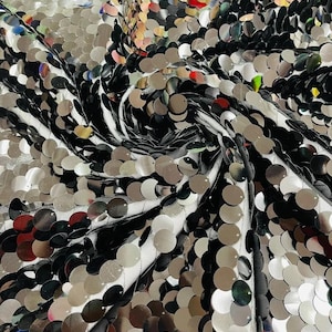 May include: A fabric featuring a dense arrangement of circular, reflective silver sequins attached to a white mesh base. The sequins are layered in a swirling pattern, creating a metallic, high-shine surface with visible light reflections.