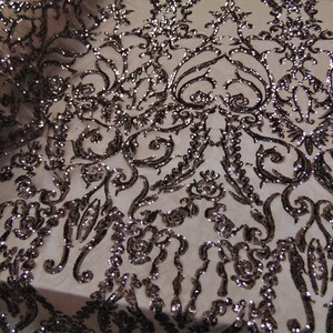 May include: A close-up of a beige fabric with a black and silver sequined pattern. The pattern is a repeating design of swirls and leaves.