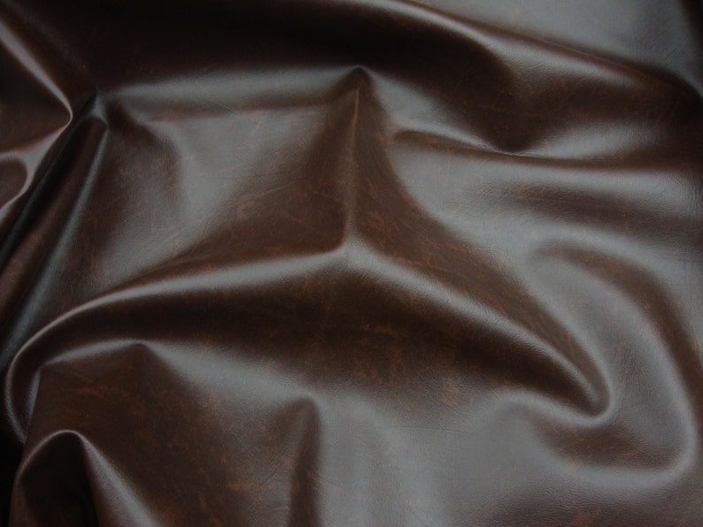 Puede incluir: Primer plano de una hoja de cuero marr&oacute;n. El cuero tiene una superficie lisa y ligeramente brillante con sutiles variaciones de color y textura. El material est&aacute; doblado y arrugado, creando sombras y reflejos.