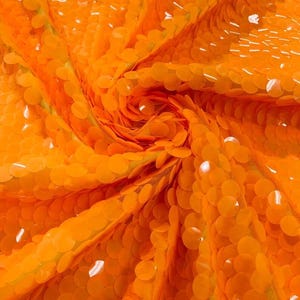 May include: A vibrant orange fabric densely covered in round, glossy sequins. The material is gathered into a central spiral, creating a dynamic, textured surface with overlapping circular discs that reflect light across the bright orange color.