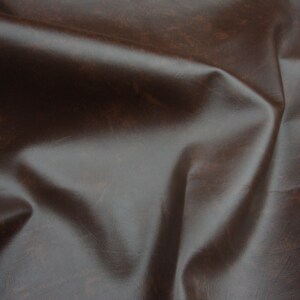 Puede incluir: Primer plano de un material de cuero marr&oacute;n oscuro con un acabado brillante. El cuero tiene un aspecto texturizado con sutiles variaciones de color y tono, y est&aacute; doblado en curvas suaves y naturales.
