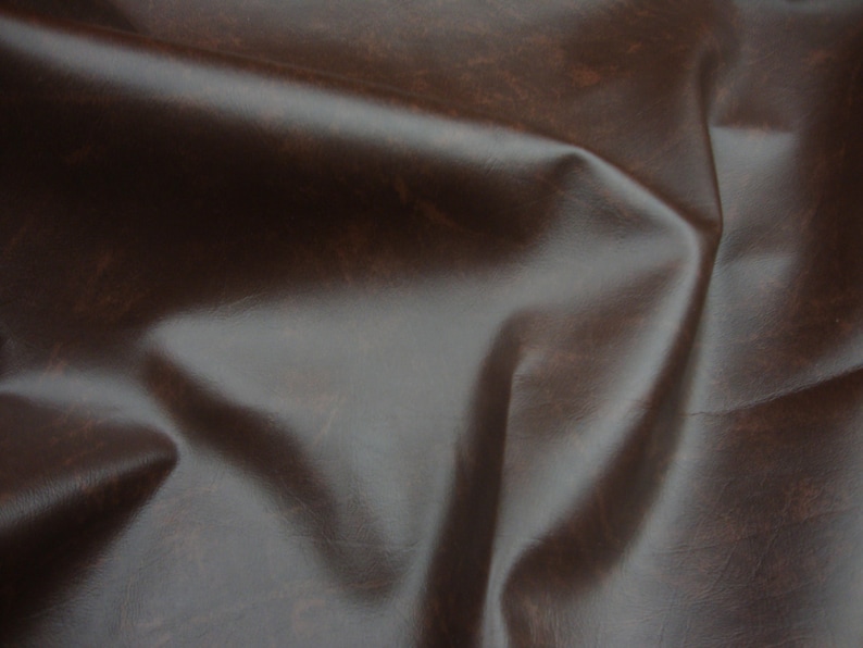 Puede incluir: Primer plano de un material de cuero marr&oacute;n oscuro con un acabado brillante. El cuero tiene un aspecto texturizado con sutiles variaciones de color y tono, y est&aacute; doblado en curvas suaves y naturales.