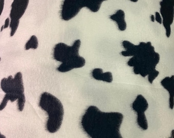 May include: Close-up of a white fabric with large, irregular black spots, resembling a cowhide pattern. The soft material is tightly woven, creating a textured surface. The black spots vary in size and shape, creating a playful and organic design.