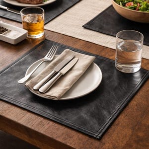 May include: A dining table setting with black placemats, white plates, silverware, and a beige linen napkin. A plate of food, a glass of water, and a glass of amber liquid are also visible. A lit candle and a wooden bowl of salad are in the background.