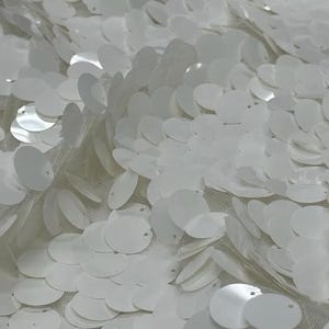 May include: Close-up of white sequin fabric. The fabric is covered in overlapping, round, white sequins. The sequins are attached to a sheer, white mesh backing. The sequins reflect light, creating a shimmering effect. This fabric is suitable for apparel and decorative projects.