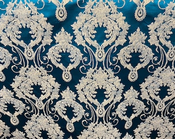 May include: Teal-colored fabric with a repeating pattern of silver-colored damask designs. The damask pattern features floral and scroll motifs, creating a luxurious and ornate appearance. The fabric has a slight sheen, enhancing the visual texture of the design.