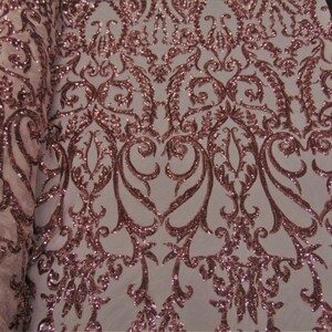 May include: A roll of fabric with a rose gold sequin design. The fabric has a white background with an ornate, symmetrical pattern. The sequins create a shimmering effect, ideal for apparel or decorative projects.