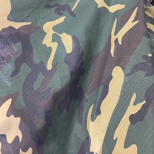 May include: Close-up of a camouflage fabric with a repeating pattern of green, brown, and tan shapes. The fabric has a textured appearance with visible diagonal lines, creating a military-inspired design.