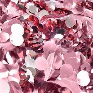 May include: Close-up of a fabric covered in shiny, round sequins. The sequins are primarily a dusty rose color, with some appearing silver. The fabric underneath is a light pink mesh.