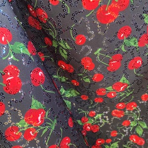 May include: Close-up of a fabric with a repeating pattern of red cherries and green leaves on a dark gray background. The cherries are embellished with sequins, adding a shimmering effect to the design. The fabric is likely used for clothing or accessories.