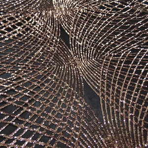 May include: A close-up of a black fabric with a rose gold sequin pattern. The sequins are arranged in a geometric design, creating a shimmering effect.