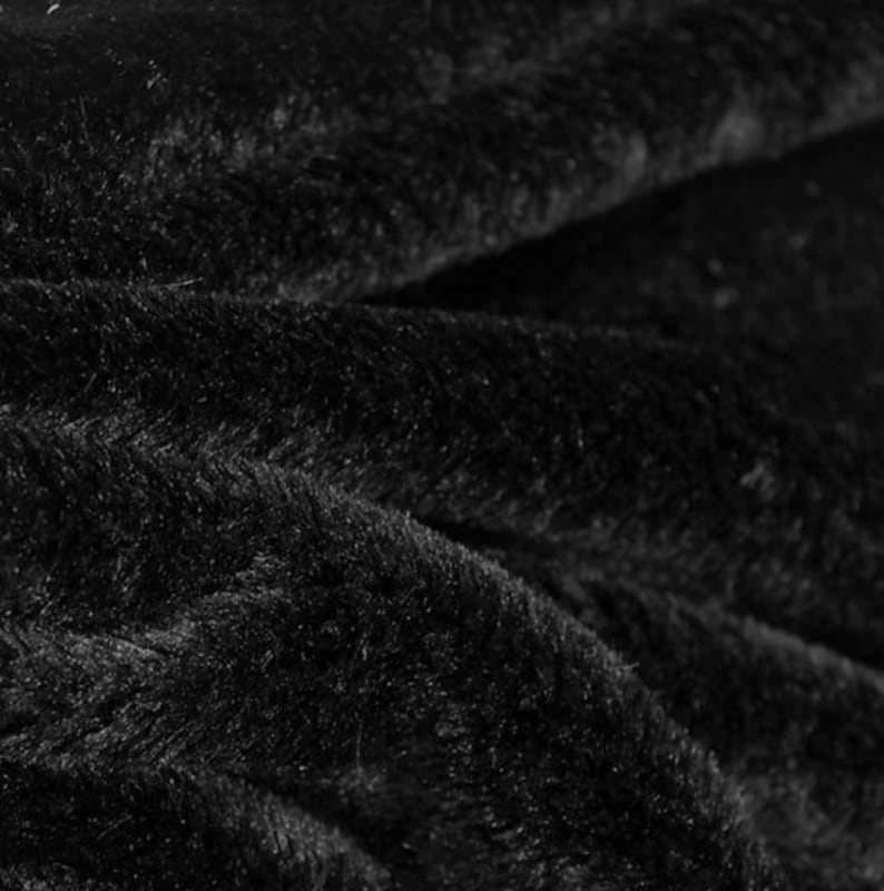 May include: Close-up of a black, plush fabric with a soft, furry texture. The material is folded, creating shadows and highlights that emphasize the depth and texture of the fabric. The overall appearance is dark and luxurious.