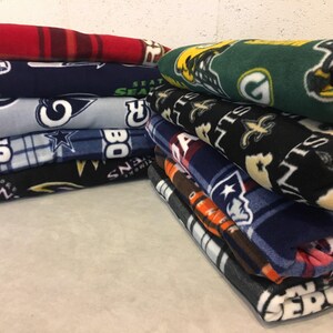 May include: A stack of fleece blankets with various NFL team logos, including the Seahawks, Cowboys, Saints, Patriots, and Jets.