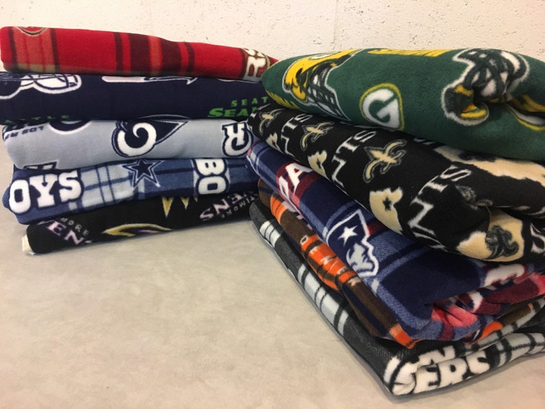 May include: A stack of fleece blankets with various NFL team logos, including the Seahawks, Cowboys, Saints, Patriots, and Jets.