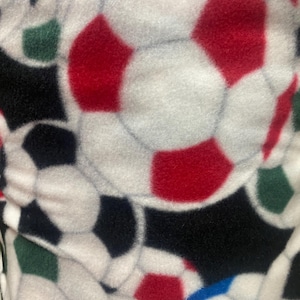May include: Close-up of a white fleece fabric patterned with soccer balls. The soccer balls have red, black, green, and blue pentagons. The background is black. The fabric is soft and plush.