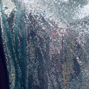 Puede incluir: Primer plano de una tela de lentejuelas plateadas con un forro azul verdoso. Las lentejuelas están dispuestas en un patrón aleatorio, creando un efecto brillante.