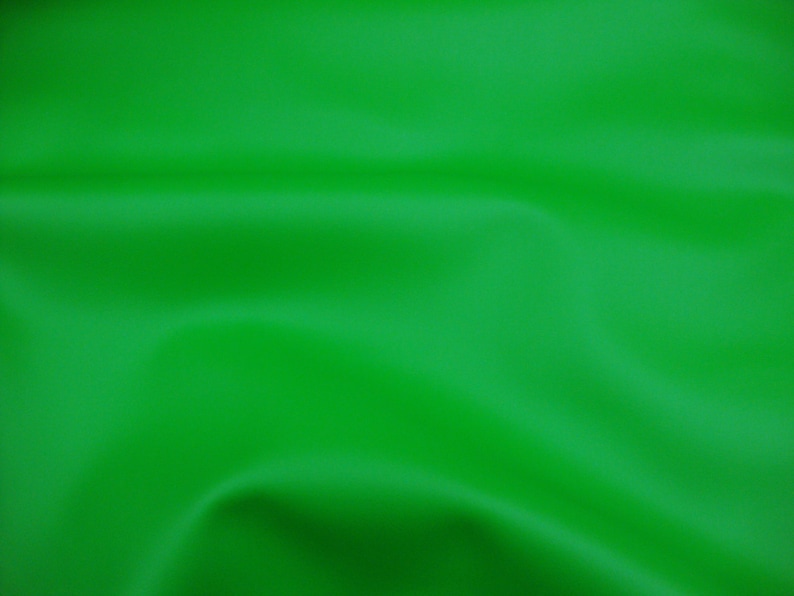 May include: A solid, vibrant green fabric with a smooth, matte texture. The material is draped in soft, flowing folds that create subtle shadows and highlights across the surface, emphasizing the consistent, bright green color throughout.