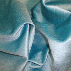 Peut inclure: Gros plan sur un tissu bleu sarcelle avec un l&eacute;ger chatoiement. Le tissu est pli&eacute; et drap&eacute;, mettant en valeur sa texture douce et ses lignes fluides.