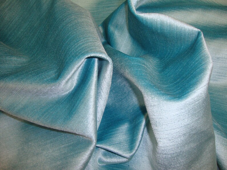 Peut inclure: Gros plan sur un tissu bleu sarcelle avec un l&eacute;ger chatoiement. Le tissu est pli&eacute; et drap&eacute;, mettant en valeur sa texture douce et ses lignes fluides.