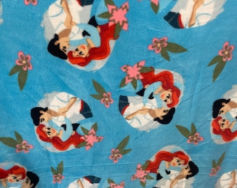 May include: A blue fabric with a repeating pattern of Ariel and Prince Eric from The Little Mermaid. The pattern includes pink flowers and green leaves.