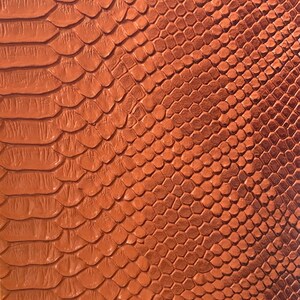 May include: Close-up of a textured, orange-brown leather material with a scale-like pattern. The design resembles snake or reptile skin, with a glossy finish. The material is likely used for fashion accessories or upholstery.