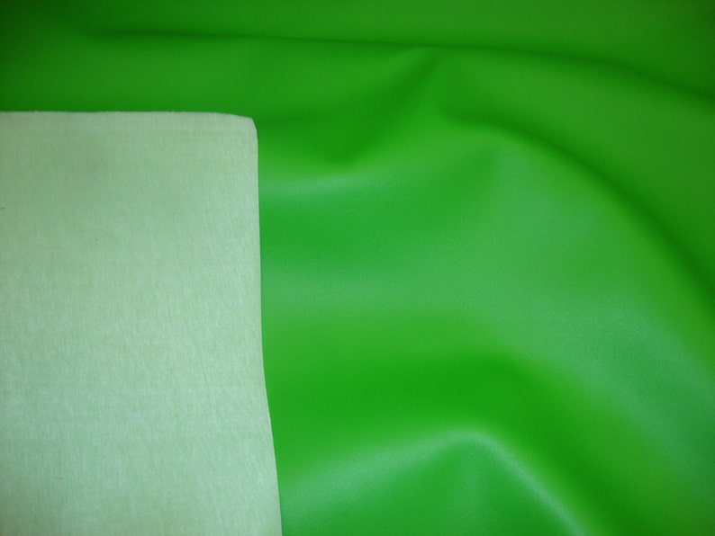 May include: A vibrant lime green fabric with a smooth, matte finish on one side and a contrasting pale, off-white felt-like backing. The material is draped to show soft folds and a flexible texture, highlighting the two distinct sides of the textile.