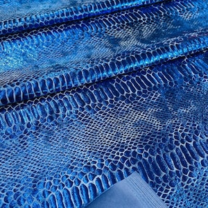 K&ouml;nnte beinhalten: Blauer, schimmernder Stoff mit Schlangenhautmuster. Der Stoff ist in mehreren Lagen gefaltet, wodurch die Textur und der Glanz des Materials sichtbar werden.
