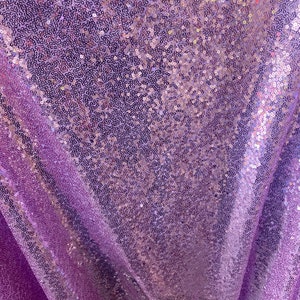 May include: Close-up of a lavender fabric covered in small, reflective sequins. The sequins catch the light, creating a shimmering effect. The fabric is draped, showing folds and texture, suitable for event decor.