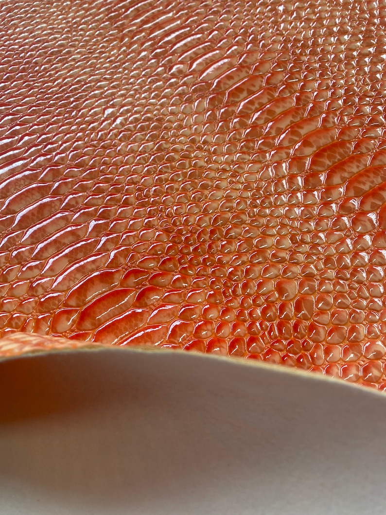 May include: Close-up of a textured, glossy material with a reptile-like pattern. The surface is a blend of orange and reddish-brown hues, with a slight sheen. The material is slightly curved, revealing a neutral-colored underside.