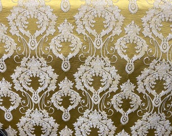 May include: Close-up of a gold-colored fabric with a white damask pattern. The fabric has a shiny, textured surface and is folded. A blue fabric with a similar pattern is visible in the background.