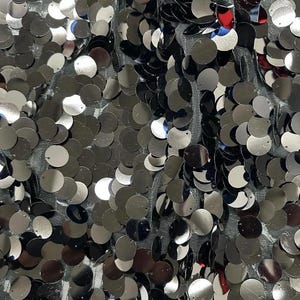 May include: Close-up of a fabric covered in shiny, silver sequins. The sequins are round and densely packed, creating a reflective surface. The fabric underneath appears to be a dark gray mesh, adding depth to the design. The overall effect is glamorous and eye-catching.