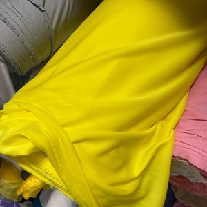 May include: Bright yellow fabric in a large roll, with soft folds and a smooth texture. Other rolls of fabric in various colors, including gray and pink, are visible in the background. The fabric appears to be a lightweight material.