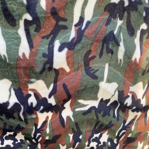 May include: Close-up of a camouflage pattern fabric. The design features irregular shapes in shades of green, brown, black, and white. The fabric has a soft, textured appearance.