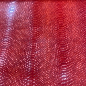 May include: A close-up of a sheet of red faux leather with a glossy finish. The material has a textured, scale-like pattern, resembling snake or reptile skin. The lighting creates highlights and shadows, enhancing the texture.