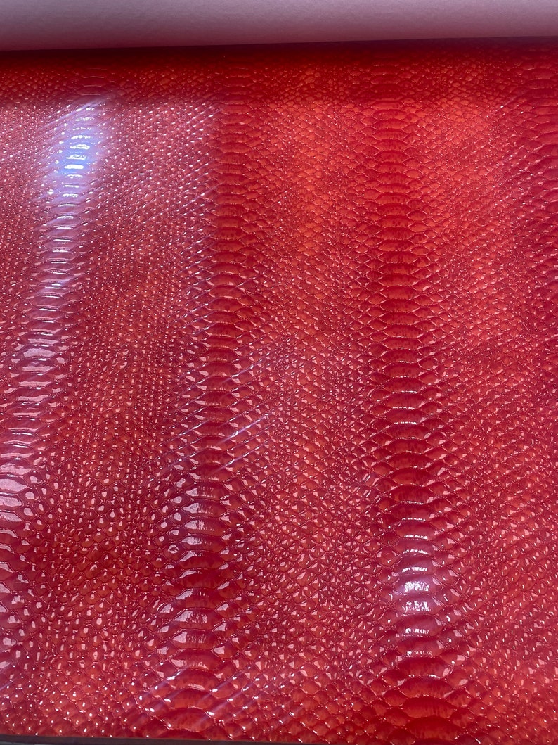May include: A close-up of a sheet of red faux leather with a glossy finish. The material has a textured, scale-like pattern, resembling snake or reptile skin. The lighting creates highlights and shadows, enhancing the texture.