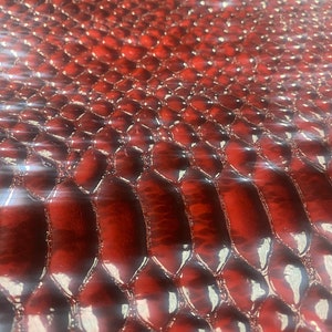 May include: Close-up of a glossy, red snakeskin pattern. The texture features a repeating scale design with a slight sheen, creating a luxurious and eye-catching appearance. The scales are slightly raised, adding depth to the surface.