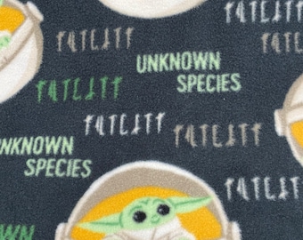 Può includere: Un tessuto nero con un motivo ripetuto di un personaggio alieno verde in un pod bianco con un interno giallo. Il testo "TATLATT" e "UNKNOWN SPECIES" è ripetuto su tutto il tessuto.