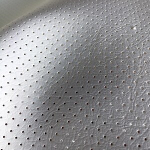 K&ouml;nnte beinhalten: Ein wei&szlig;es Material mit einer strukturierten, lederartigen Oberfl&auml;che, das ein gleichm&auml;&szlig;iges Muster aus kleinen, gleichm&auml;&szlig;ig verteilten kreisf&ouml;rmigen Perforationen aufweist. Das Material wird von oben links beleuchtet, was einen sanften Schattenverlauf erzeugt.