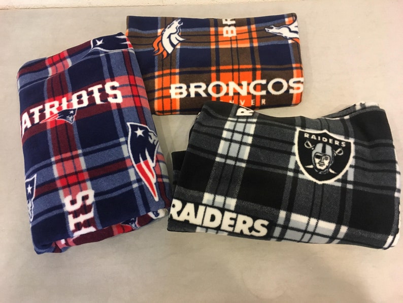 May include: Three fleece blankets with NFL team logos. The blankets are folded and feature the logos of the New England Patriots, Denver Broncos, and Las Vegas Raiders.