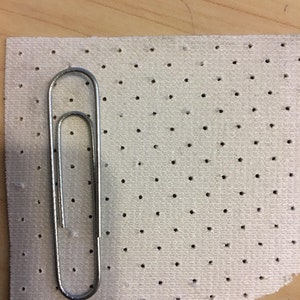 K&ouml;nnte beinhalten: Eine silberne Metall-B&uuml;roklammer liegt auf einem rechteckigen St&uuml;ck wei&szlig;em Stoff, der ein gleichm&auml;&szlig;iges Raster aus kleinen, gleichm&auml;&szlig;ig verteilten kreisf&ouml;rmigen Perforationen aufweist und einen klaren visuellen Ma&szlig;stab f&uuml;r das Stoffmuster bietet.