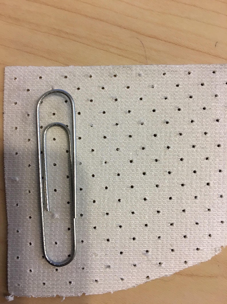 K&ouml;nnte beinhalten: Eine silberne Metall-B&uuml;roklammer liegt auf einem rechteckigen St&uuml;ck wei&szlig;em Stoff, der ein gleichm&auml;&szlig;iges Raster aus kleinen, gleichm&auml;&szlig;ig verteilten kreisf&ouml;rmigen Perforationen aufweist und einen klaren visuellen Ma&szlig;stab f&uuml;r das Stoffmuster bietet.