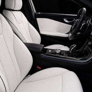 May include: The interior of a vehicle featuring white perforated leather seats with black piping. The center console is finished in a glossy black material with metallic accents, a gear shifter, and a circular control dial near the armrest.