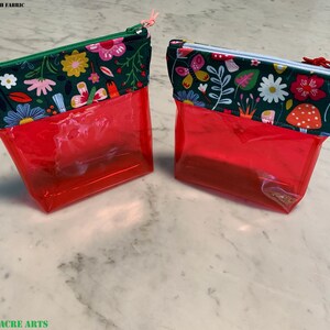 Puede incluir: Dos bolsas transparentes rojas con cremallera con tapas de tela floral. La tela de la bolsa de la izquierda presenta una variedad de flores y hojas en rosa, amarillo, azul y verde sobre un fondo verde oscuro. La tela de la bolsa de la derecha presenta una variedad de flores, hojas y hongos en rosa, amarillo, azul y verde sobre un fondo verde oscuro. El texto "PRODUCT MADE WITH FABRIC" es visible en la parte superior de la imagen.