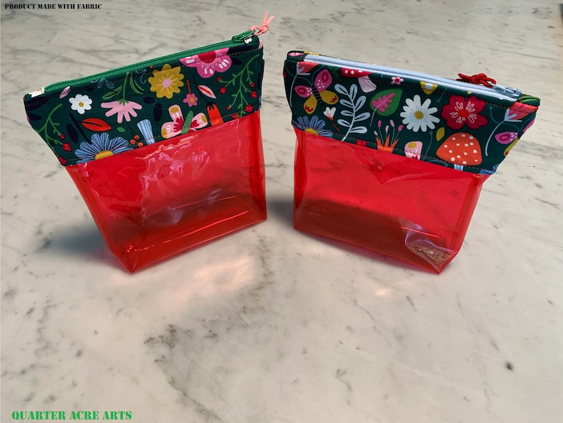 Puede incluir: Dos bolsas transparentes rojas con cremallera con tapas de tela floral. La tela de la bolsa de la izquierda presenta una variedad de flores y hojas en rosa, amarillo, azul y verde sobre un fondo verde oscuro. La tela de la bolsa de la derecha presenta una variedad de flores, hojas y hongos en rosa, amarillo, azul y verde sobre un fondo verde oscuro. El texto "PRODUCT MADE WITH FABRIC" es visible en la parte superior de la imagen.