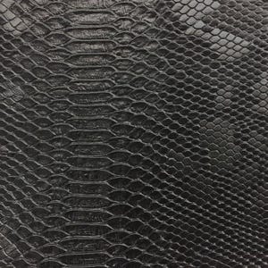 May include: Close-up of a black, textured material with a snakeskin pattern. The surface has a repeating scale design, with ridges and grooves creating a realistic reptile skin appearance. The color is a deep, solid black.